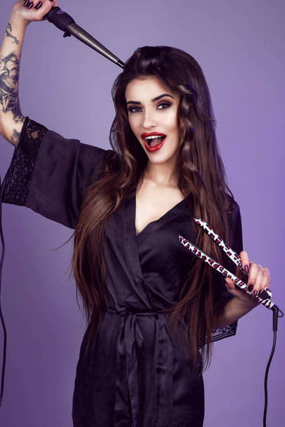 Beauty portrait of young gorgeous laughing model with long brown hair wearing black silk peignoir holding curling wand and hair straightener - Photo, Image