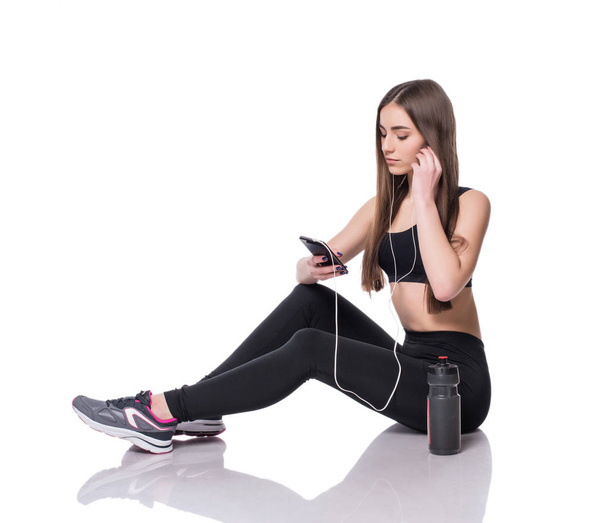 Portrait of a young athlete woman listening to music with earphones over white background. Attractive fitness girl chatting on smartphone ion studio. - Photo, Image