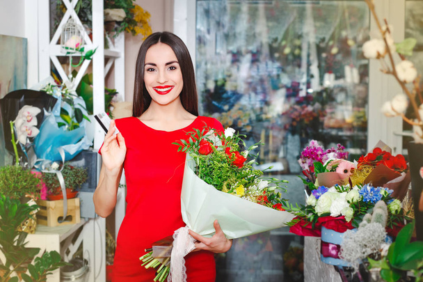 Beautiful brunette in red dress buys a bouquet of flowers in a flower shop - Фото, изображение