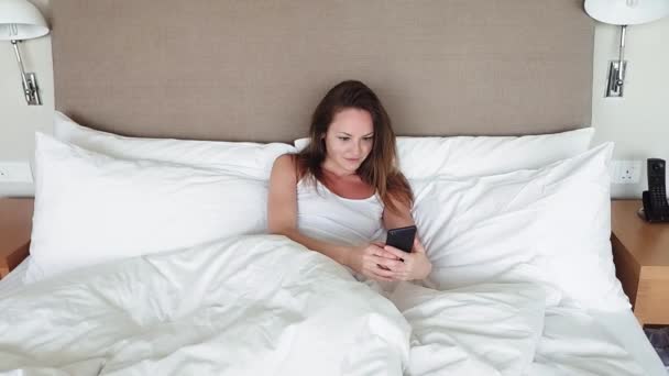 Mulher deitada na cama verificando mensagens no telefone móvel - Filmagem, Vídeo