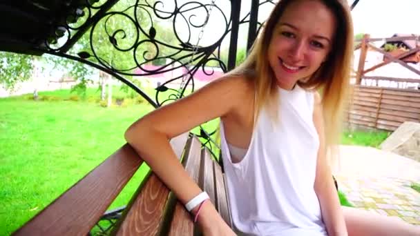 Aranyos fiatal lány ült, és ringató a padon, látszó-on fényképezőgép mosolyogva és szabadidő szabadban nap portréja. - Felvétel, videó