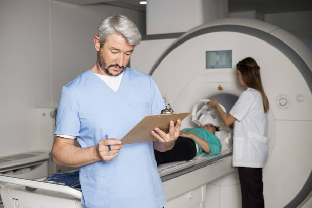 Doctor Reading Clipboard With Colleague Preparing Patient For CT - Photo, Image