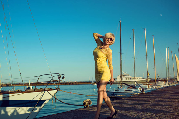 Ritratto di splendida bionda di mezza età che sorride indossando un mini abito di pizzo giallo aderente e occhiali da sole che ballano sul molo dello yacht
.  - Foto, immagini