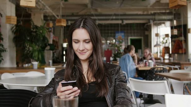 Mujer joven con estilo utiliza un teléfono inteligente navegar por Internet sentado en una mesa en la cafetería moderna, sonriendo. 4K
 - Foto, imagen