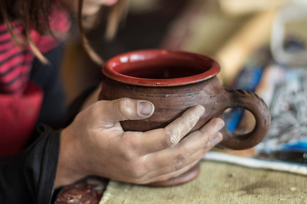 Young girl dirty in clay hands holding a ceramic mug that she made herself on the potters wheel in pottery school. Selective focus. - Foto, imagen