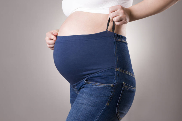 Pregnant woman in blue jeans for pregnant women on gray background - 写真・画像