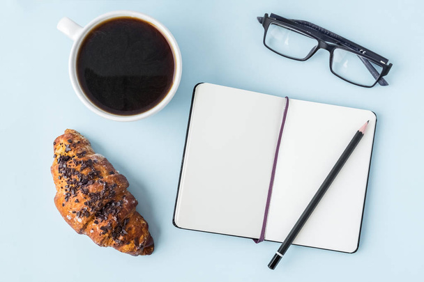 Office blue table with blank notepad, pencil, glasses, fresh croissant and coffee cup. Copy space for text. Flat lay. Top view. - Foto, immagini