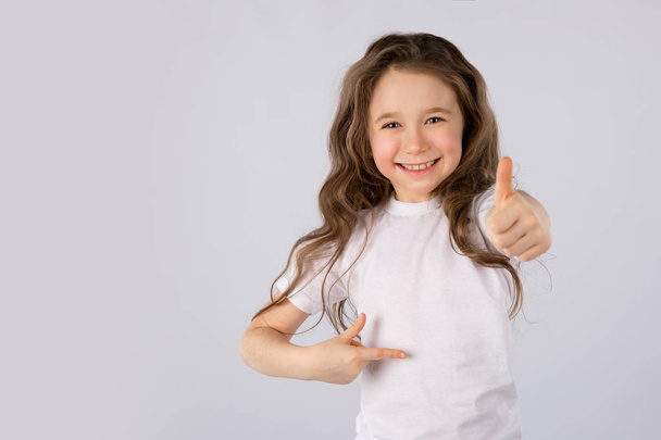 Little girl showing thumbs up gesture in a white T-shirt on white background. - Photo, image