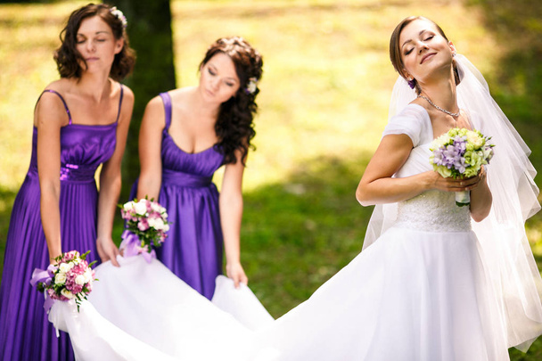 Bride enjoys a sunshine while bridesmaids hold her dress on the  - Фото, зображення
