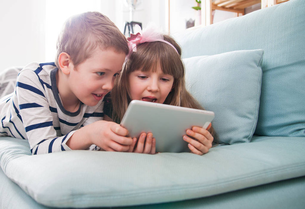 Happy siblings lying on sofa at home and playing with tablet together - Фото, зображення