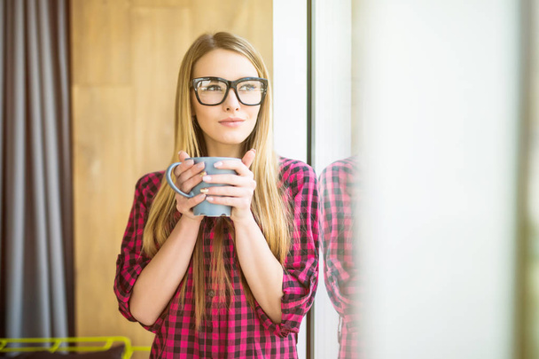 Young freelance lady drinking coffee in casual clothes and looking through a window. Morning. - 写真・画像