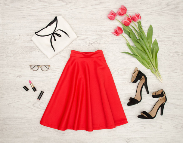 Divat fogalmát. Piros szoknya, blúz, napszemüveg, rúzs, fekete cipő és rózsaszín tulipánok. Felülnézet, világos fa háttér - Fotó, kép