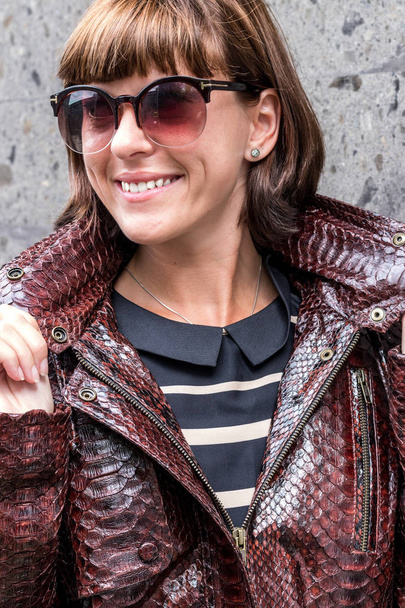 Mujer joven hermosa y de moda en una larga chaqueta de piel de serpiente de pitón de lujo posando cerca de la pared gris
. - Foto, Imagen