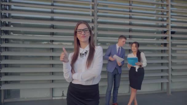 Gyönyörű fiatal üzleti nő mosolyogva és azt mutatja, gesztus a remek és áll a háttérben a kollégák akik megvitassák munkafolyamatok semleges színek. - Felvétel, videó