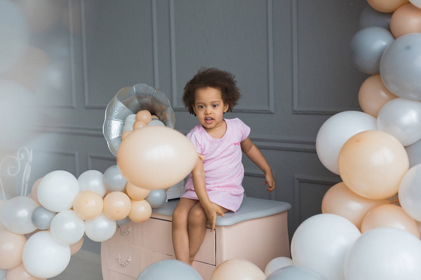 Black little girl sits and plays with colorful balloons in a studio - 写真・画像
