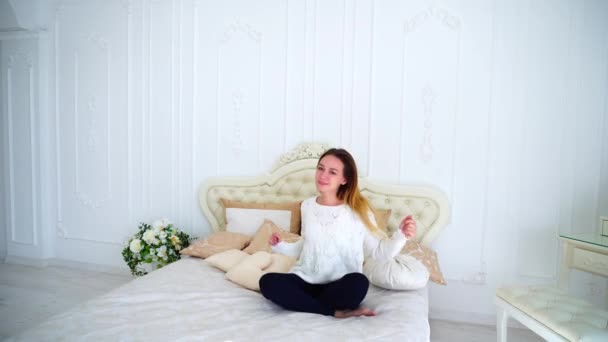 Hyvännäköinen nuori nainen onnellinen ja nauraa kameralle, istuu kauniilla tanssisängyllä valkoisessa makuuhuoneessa
. - Materiaali, video
