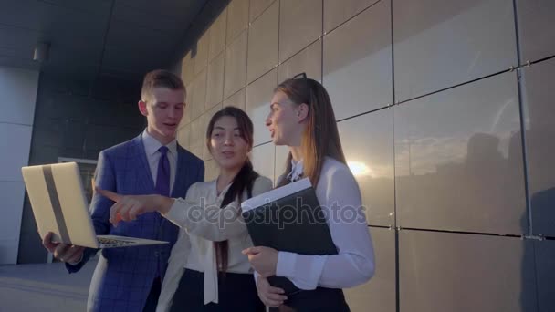 Τρεις όμορφες νέοι, σπουδαστές κρατούν στα χέρια Laptop και φακέλους, μιλάμε για το φόντο του επιχειρηματικού κέντρου σε εξωτερικούς χώρους σε ουδέτερα χρώματα. - Πλάνα, βίντεο