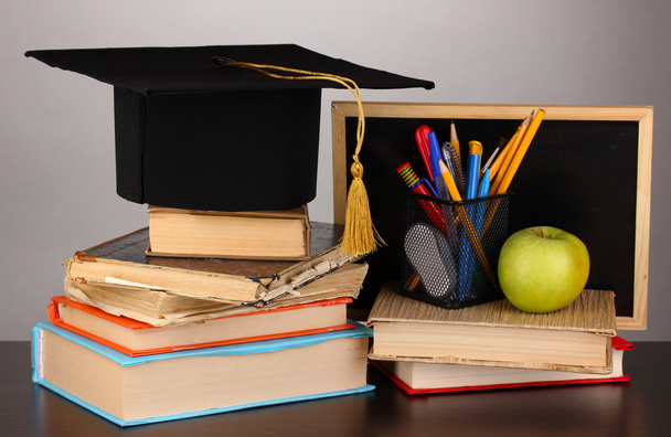 Books and magister cap against school board on wooden table on grey background - Foto, afbeelding