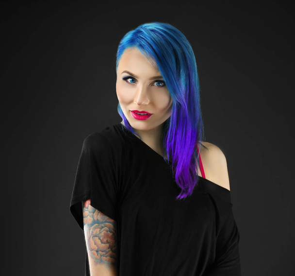 Όμορφη νεαρή γυναίκα με το κομψό χρώμα μαλλιών και τατουάζ σε σκούρο φόντο - Φωτογραφία, εικόνα