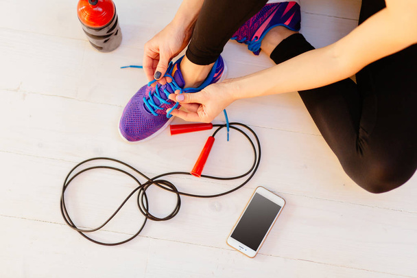 Top view of woman sitting on white wooden floor and tying her sport shoes. Skipping rope, bottle of water and mobile phone on the white wooden background. Sport, diet and healthy life concept. - Fotó, kép