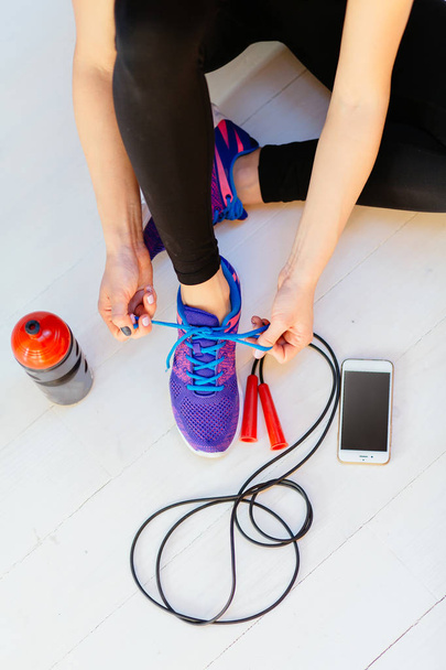 Top view of woman sitting on white wooden floor and tying her sport shoes. Skipping rope, bottle of water and mobile phone on the white wooden background. Sport, diet and healthy life concept. - Photo, Image