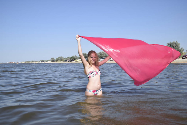 Blond girl in a bikini standing on the beach and holding a red silk handkerchief. Beautiful young woman in a colorful bikini on sea background. - Фото, зображення