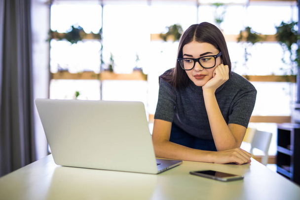 fröhliche junge schöne Frau mit Brille, die lächelnd auf ihren Laptop blickt, während sie an ihrem Arbeitsplatz sitzt - Foto, Bild