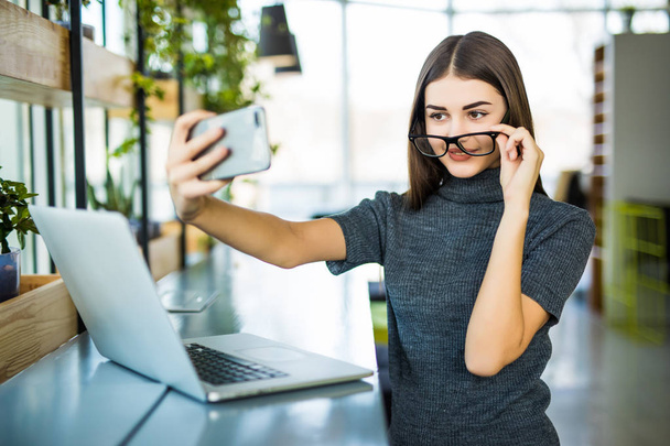 junge Frau macht ein Selfie im Büro, während sie mit Laptop sitzt. - Foto, Bild