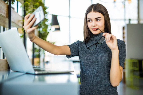 Nuori nainen ottaa selfie toimistossa lasit istuessaan kannettavan tietokoneen kanssa
. - Valokuva, kuva