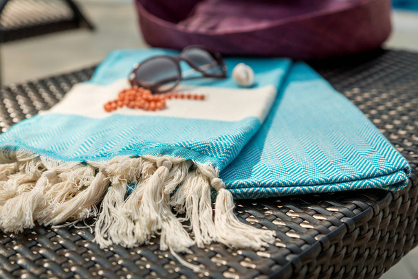 The Concept of flat lay summer accessories of white turquoise color Turkish towel / peshtemal, sunglasses, seashell, orange necklace and crimson / redviolet color straw hat on rattan lounger . - Photo, Image