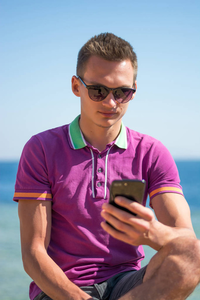 Mies aurinkolaseissa istuu kännykkä kädessään lähellä merta.
 - Valokuva, kuva