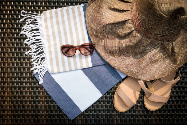 The concept of flatlay summer accessories of white, blue and beige Turkish towel, sunglasses, beige leather ladies sandals and straw hat on a rattan lounger. - Photo, Image