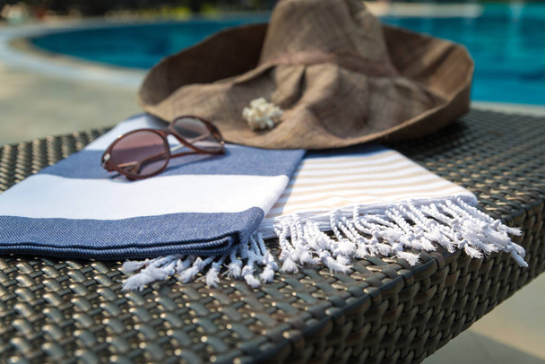 Concept of summer accessories close-up of white, blue and beige Turkish towel, sunglasses and straw hat on rattan lounger with blue swimming pool as background. - Photo, image