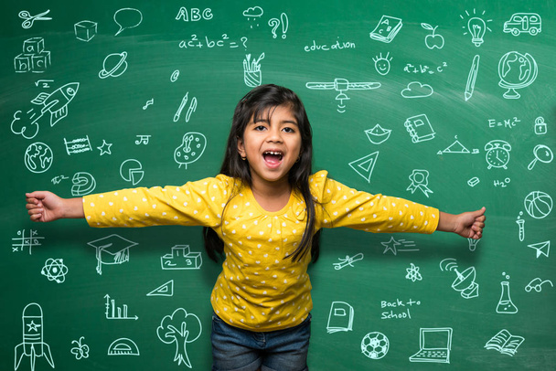 Indian school kid girl in hand stretched pose over green chalkboard or chalk board background, indian boy standing over green chalkboard background in the school with doodles -  back to school concept - 写真・画像