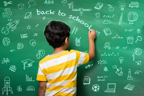 Indian school kid in hand stretched pose over green chalkboard or chalk board background, indian boy standing over green chalkboard background in the school with doodles -  back to school concept - Photo, Image