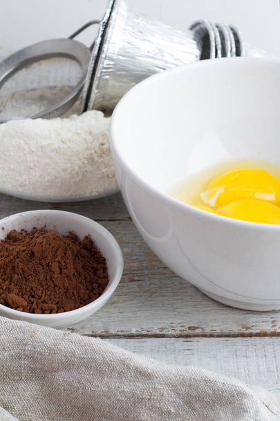 Kom met meel, eieren, cacao en melk op een witte achtergrond - Foto, afbeelding