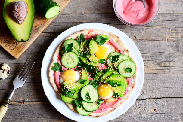 Tortilla piled high with fried quail eggs, avocado, cucumbers, sesame and parsley and lathered with beetroot hummus. Half avocado, beetroot hummus in bowl, fork on vintage wooden background. Top view - Foto, Bild