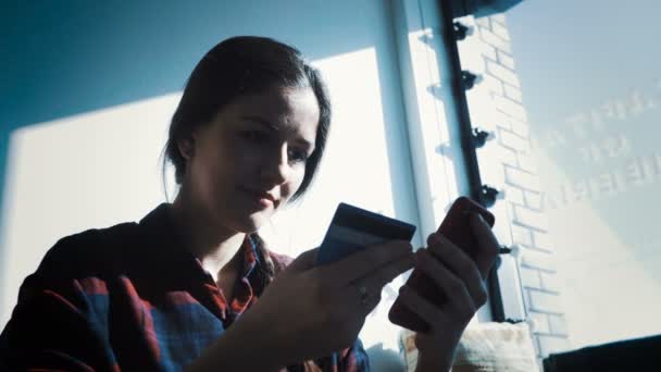 Smiling young woman sitting at table using touchpad and holding credit card in her hand - Felvétel, videó