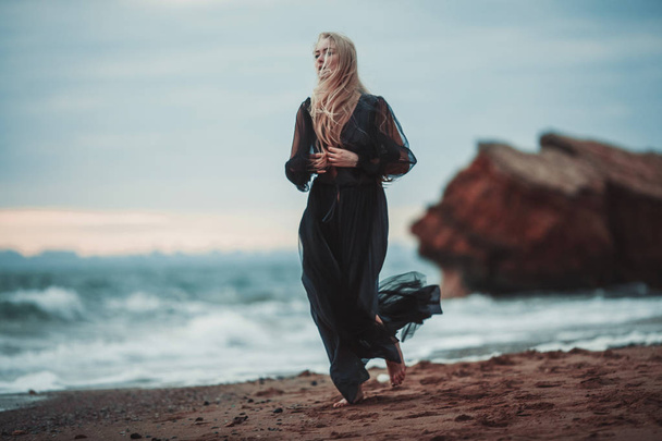 junge Frau im langen transparenten schwarzen Kleid zwischen Strand und Meer. - Foto, Bild