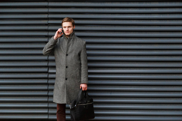 Jonge vertrouwen zakenman in een grijze jas en de werkmap in zijn hand gaat op mobiele telefoon tegen muur van modern gebouw - Foto, afbeelding