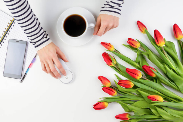Woman hands with cup of coffee and computer mouse. Mobile phone, paper notebook and beautiful flowers on white table - 写真・画像