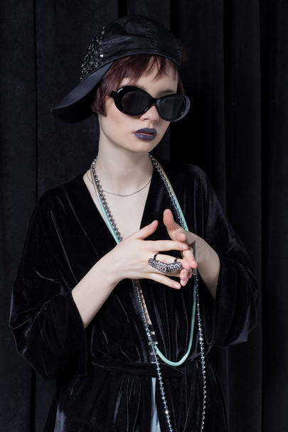 fashion shot of a beautiful young girl with a cheeky short hair in a hipster style in a black velvet jacket in Kupka Chesney and sunglasses in a studio on a black background - Photo, image