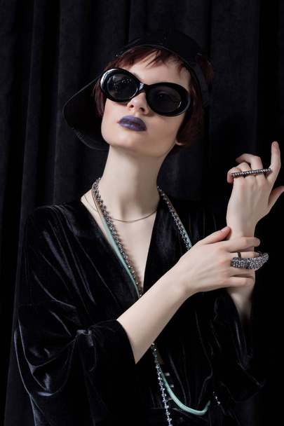 fashion shot of a beautiful young girl with a cheeky short hair in a hipster style in a black velvet jacket in Kupka Chesney and sunglasses in a studio on a black background - Foto, immagini