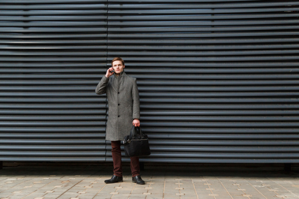 Magabiztos fiatal üzletember egy szürke kabátot és a táskáját a kezében mobiltelefon modern épület falnak beszél. - Fotó, kép