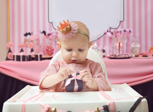 Cute little birthday girl with present sitting on high chair in room decorated for party - Foto, imagen