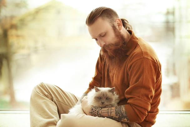 Young bearded man with fluffy cat sitting on floor - Фото, изображение