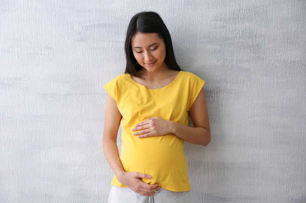 Pregnant Asian woman standing near light wall - Фото, изображение