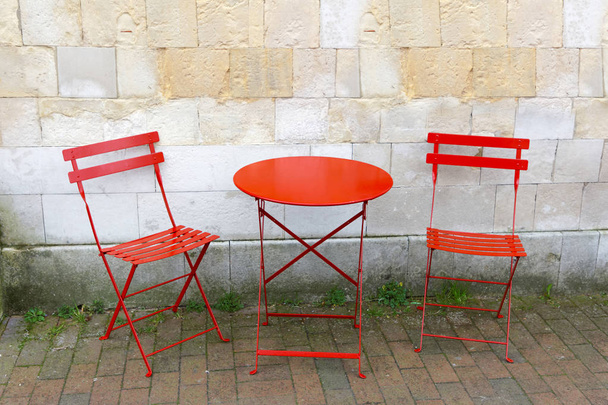 Duas cadeiras e uma mesa vermelha na frente de uma parede de pedra cinza
 - Foto, Imagem