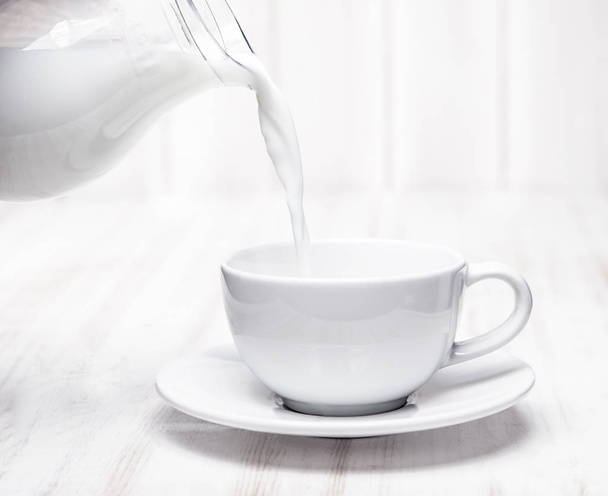 Milk is poured into a cup of pitcher on white background wooden  - Фото, зображення