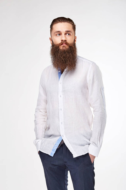 Young bearded man posing in studio in trendy linen white shirt and blue pants - 写真・画像